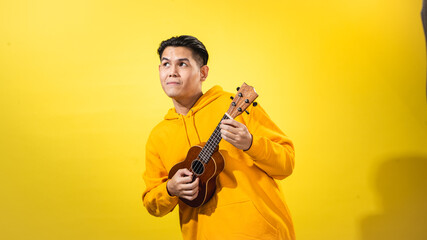 Asian man in yellow hoodie enthusiastically playing a small ukulele against a vivid yellow...