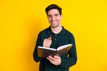 Handsome young man with glasses holding a notebook and pen, smiling confidently against a vibrant yellow background