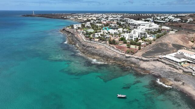 Panoramaflug entlang der K&uuml;ste von Playa Blanca, Lanzarote: t&uuml;rkisfarbenes Meer, schwarze Felsen, Hotels, Palmen und vulkanische Landschaften aus der Vogelperspektive.