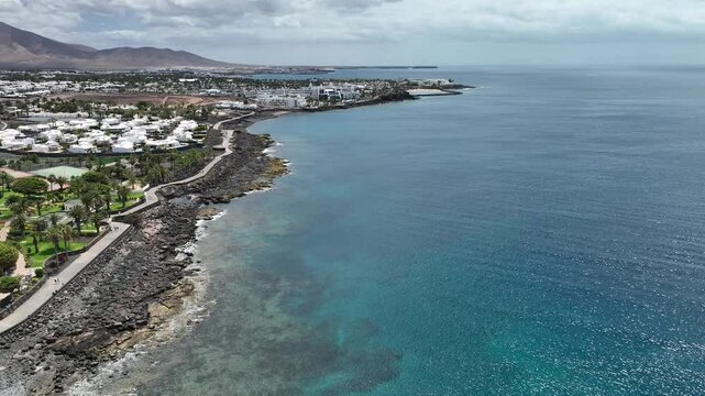 Panoramaflug entlang der K&uuml;ste von Playa Blanca, Lanzarote: t&uuml;rkisfarbenes Meer, schwarze Felsen, Hotels, Palmen und vulkanische Landschaften aus der Vogelperspektive.