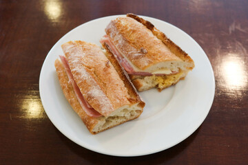 close up of a Baguette bread sandwich with ham and cheese cut in two half on a white plate with copy space