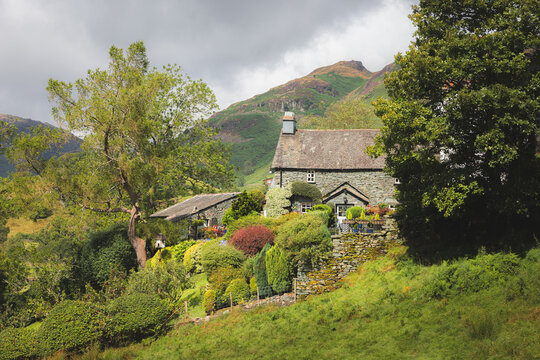 A quaint stone cottage in Langdale, near Ambleside, is surrounded by a serene garden and greenery, set against the picturesque Lake District mountains.