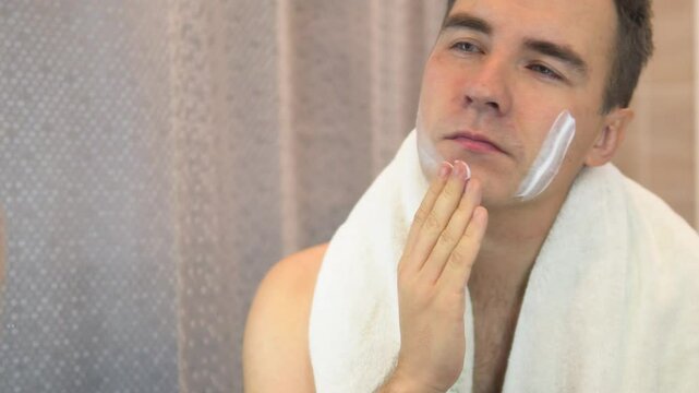 A young guy with a white jersey applies shaving foam to his face and looks in the mirror. Bathroom with daylight. Male skin care.