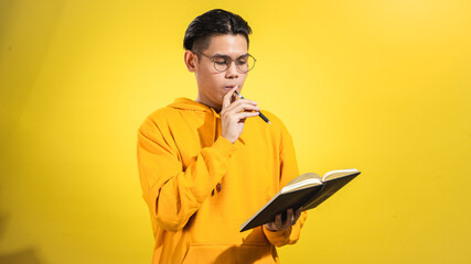 Thoughtful young Asian man in yellow hoodie wearing glasses, holding a pen and notebook while reading and thinking, standing against solid yellow background. Studio concept of planning and learning