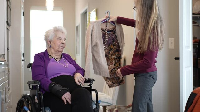A mature woman seated in a wheelchair engages with a caregiver who holds up clothing options in a well-lit hallway at home. The interaction suggests companionship and care.