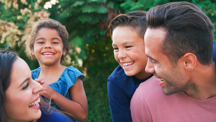 Portrait Of Smiling Hispanic Family With Parents Giving Children Piggyback Rides In Garden At Home