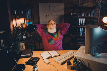 Senior man in cozy sweater enjoys a relaxed moment, working on computer technology in a home office environment under warm light