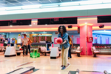 Fototapeta premium Cheerful young woman throwing a bowling ball, enjoying a friendly game with friends in a vibrant bowling alley