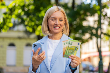 Caucasian mature businesswoman standing on city street showing earnings winning at betting cash money with mobile phone in hands. Middle-aged woman in a suit outdoors with smartphone and pack euro.