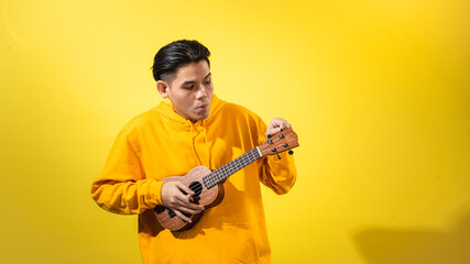 Asian man in yellow hoodie enthusiastically playing a small ukulele against a vivid yellow background. Expressive musical portrait with happy mood and creative, artistic personality