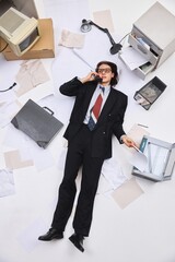 Businessman wearing in stylish formal suit lying on back holding paper and phone surrounded by...
