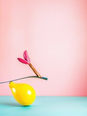 A yellow balloon balanced intricately on a thin dark rod with a pink flower dart against a soft pastel pink background and light teal surface below