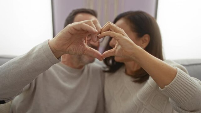 Couple forming heart shape with hands in cozy living room, symbolizing love and togetherness, middle-aged hispanic man and woman sharing intimate moment indoors in home setting.