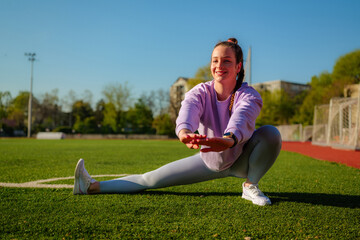 Young woman wearing sportswear performing leg stretches on a green turf field, enjoying a sunny day...