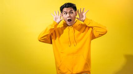 Curious young Asian man in yellow hoodie holding his hand to ear as if listening, with surprised...