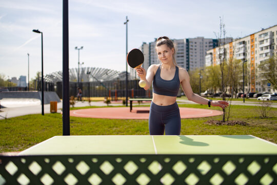 A woman is playing table tennis on the street. She's wearing a tracksuit and holding a racket. Residential buildings and trees are visible in the background.