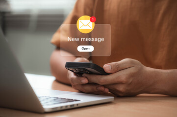 Young person holding smartphone receiving new email message alert, sitting at desk with laptop in casual outfit, representing real time digital communication and modern email marketing lifestyle
