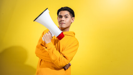 Confident young Asian man in yellow hoodie holding a red and white megaphone with calm expression, standing against a solid yellow background. Studio concept of message delivery or leadership voice