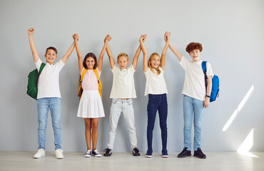 Obraz premium Happy smiling group of school kids in casual clothes standing together in a row holding hands up isolated on a gray wall background. Schoolchildren with backpacks looking cheerful at camera.