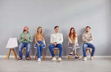 Naklejka premium Young people men and women in casual clothes sitting on chairs in row waiting for interview invitation turn in office. Job candidates seekers sitting on gray wall background. Recruitment concept.