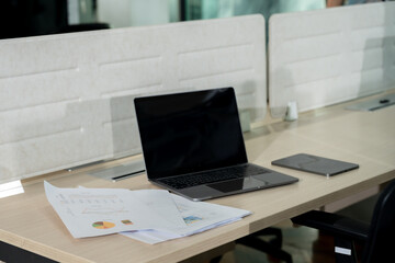 Modern open office workspace with unoccupied wooden desk showing digital tablet, closed laptop, printed business charts, and partition divider indicating ongoing collaborative office work environment