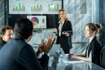 Businesswoman presenting during office meeting holding stylus and tablet while smiling team listening with interest teamwork communication collaboration on data charts displayed on presentation screen