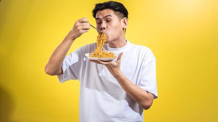 Asian man in a white t-shirt enthusiastically eating noodles from a plate with a fork, looking to...