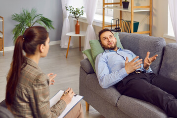 Adult male patient talking to counseling psychologist at therapy session. Young man lying on sofa, sharing thoughts with female therapist, expressing doubt, asking for advice. Psychotherapy concept