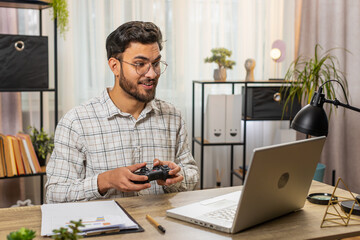 Good-looking excited Hispanic businessman celebrating victory in video online game on laptop spending leisure time at home office desk. Happy freelancer playing video games holding controller