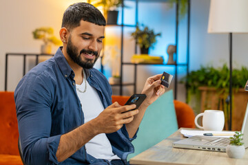 Young Indian man freelancer using credit bank card and smartphone while transferring money, online...