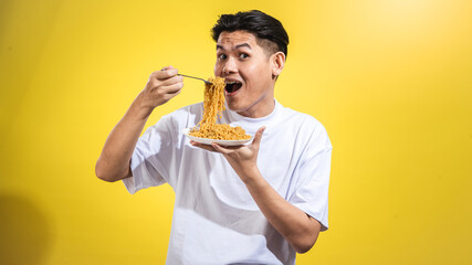 Asian man in a white t-shirt enthusiastically eating noodles from a plate with a fork, looking to...