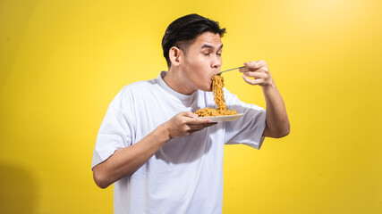 Asian man in a white t-shirt enthusiastically eating noodles from a plate with a fork, looking to...