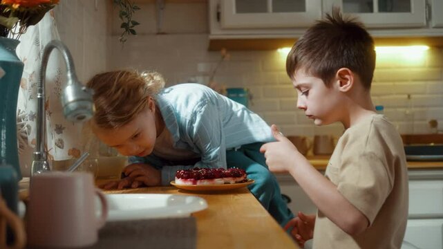 Two little children in the kitchen are sweetly eating birthday cake with their fingers and having fun, and they smear each other in the face with whipped cream.