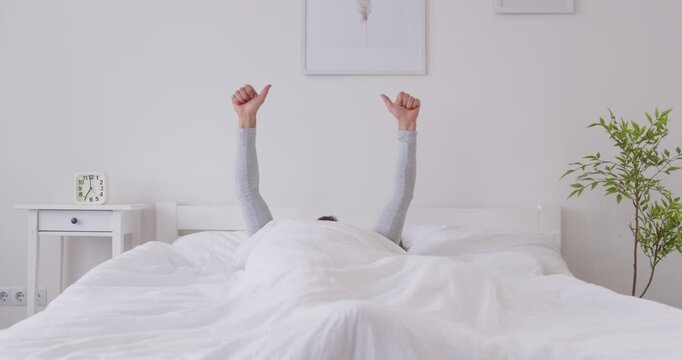 Male person lying in bed under white cozy blanket stretching his arms and showing thumb up sign in modern bedroom. Relaxed sleepy man waking up in the morning with good mood. 4k video.