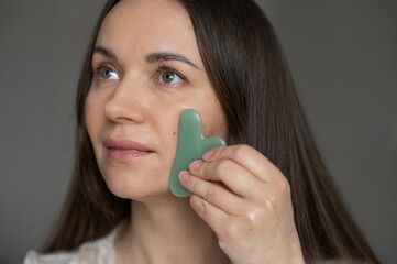 Young woman using a green gua sha scraper for facial massage, improving skin circulation and...