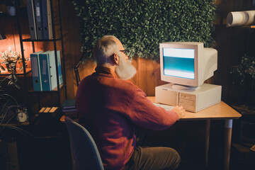 Senior professional working on a vintage computer in a cozy home office surrounded by greenery...