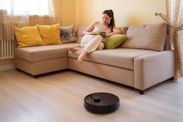 A robot vacuum cleaner washes the floor while a young woman reads a book sitting on the sofa. Smart home, automation and modern technologies in everyday life