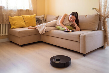 A robot vacuum cleaner washes the floor while a young woman reads a book sitting on the sofa. Smart...