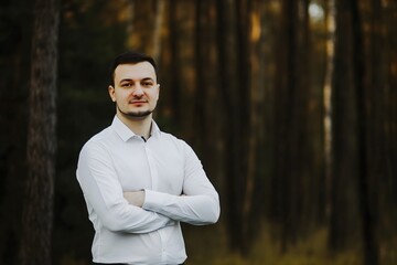 Confident young professional man in a classic white shirt stands with arms crossed, smiling for a business headshot in a natural autumn forest setting