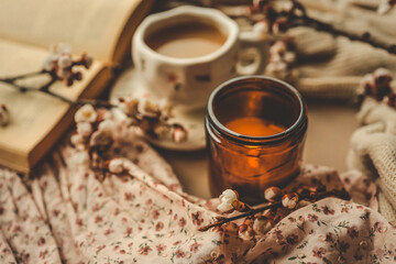 Warm cozy scene with a burning candle, a cup of tea, blooming flowers, an open book, and a floral fabric, creating a peaceful spring atmosphere