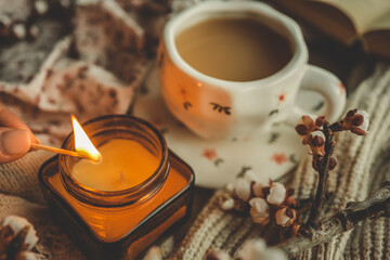 Hand lighting a candle next to a cup of tea in a floral mug, with spring blossoms and cozy textures in a warm, relaxing atmosphere