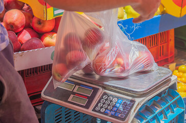 Transparent plastic bags with ripe fruits of red peaches, apples, which seller at market lays out on surface of measuring part of electronic floor scale. Trade, purchase of farm products for nutrition