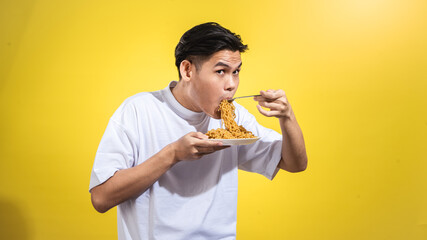 Asian man in a white t-shirt enthusiastically eating noodles from a plate with a fork, looking to...