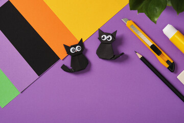 Paper origami black cats with puffy eyes on purple background. Around crafting supplies, paper...
