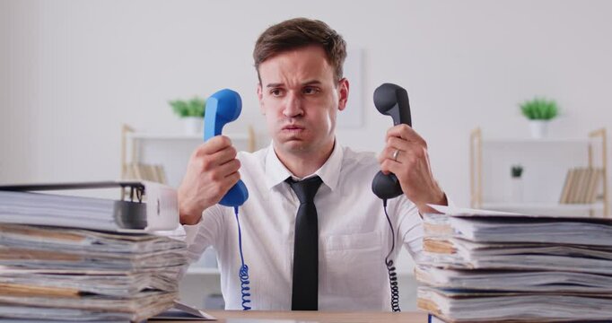 Tired upset young business man company employee holding different handsets sitting at desk in office with a pile of documents. Overwork and multitasking concept. 4k video. Slow motion video.
