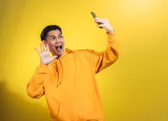 Cheerful young Asian man in yellow hoodie smiling and waving at smartphone while taking selfie against bright yellow background. Playful studio shot capturing modern communication and joy