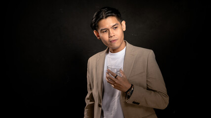 Asian man in beige blazer and white t-shirt holding a transparent glass, looking away with contemplative expression in studio with dark background, perfect for lifestyle or branding visuals
