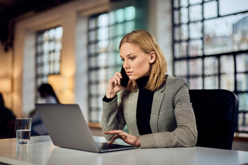 Fototapeta premium Focused Professional Woman Talking on Phone While Using Laptop at Office Desk