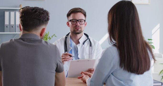 Doctor consulting young family couple planning children and visiting doctor clinic. Explaining details of IVF treatment, patients listening to male gynecologist at health center or modern hospital