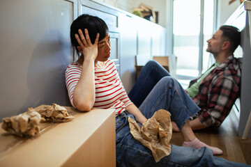 Stressed couple sitting on floor surrounded by moving boxes in new home